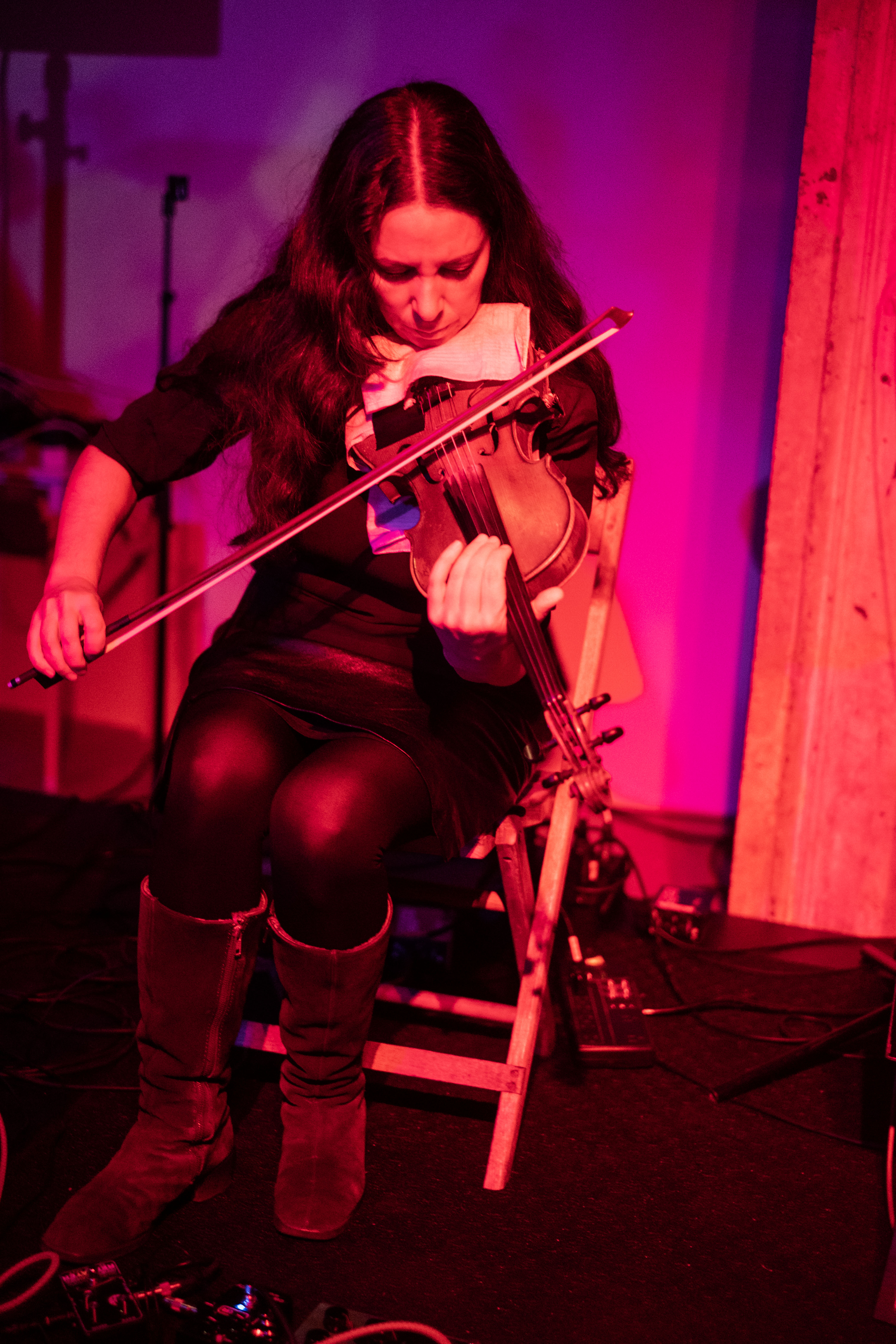 A seated figure plays the violin on stage with red and purple lights cast onto them.
