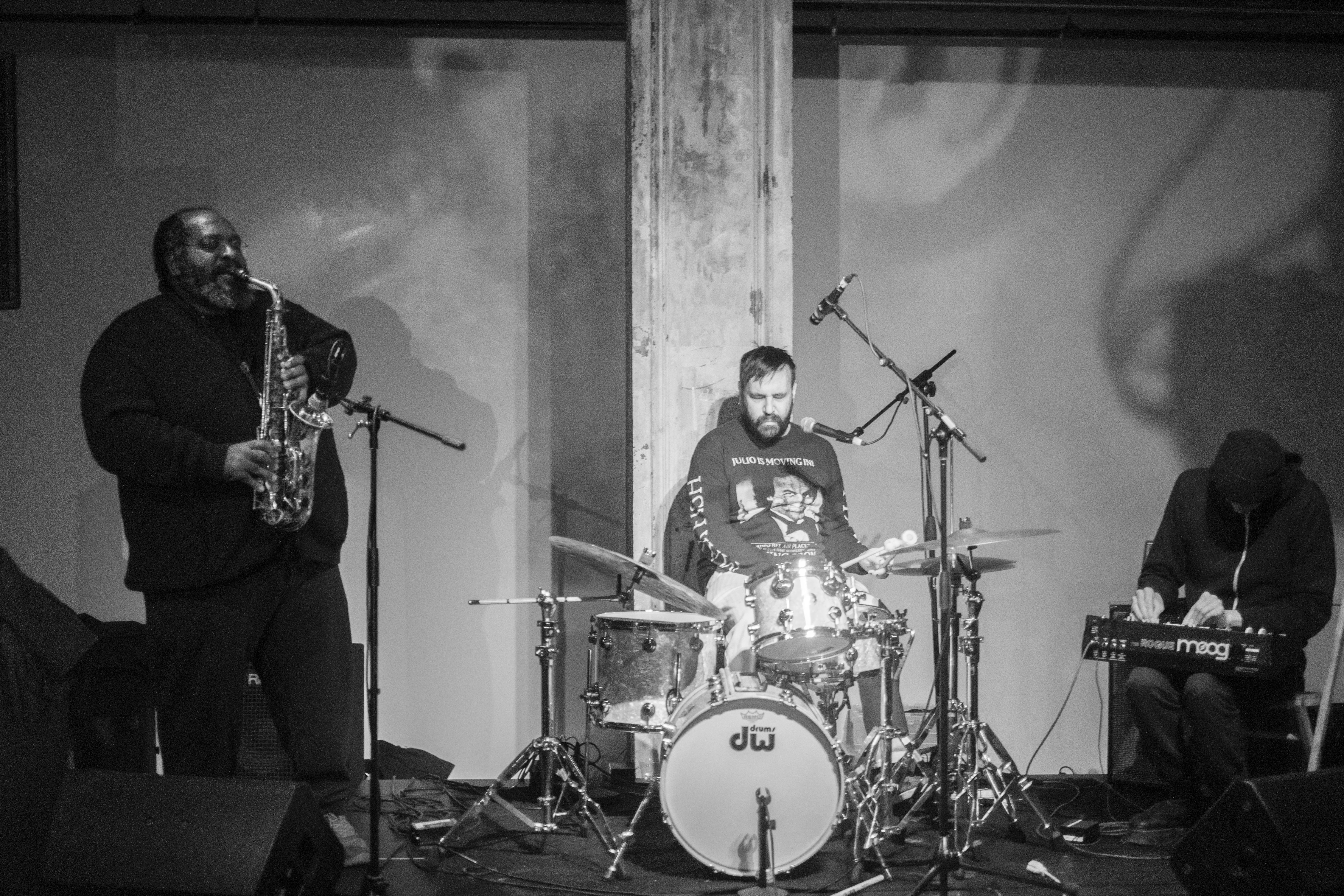 Black-and-white image of three figures performing in a band on stage, the right figure plays the saxophone, the middle figure plays the drums, and the right figure plays a synthesizer.
