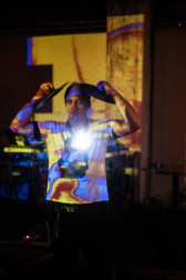 A figure in a white graphic T-shirt has their eyes closed, while they stand and hold two cymbals over their head. Yellow and blue projections are cast onto them on stage.