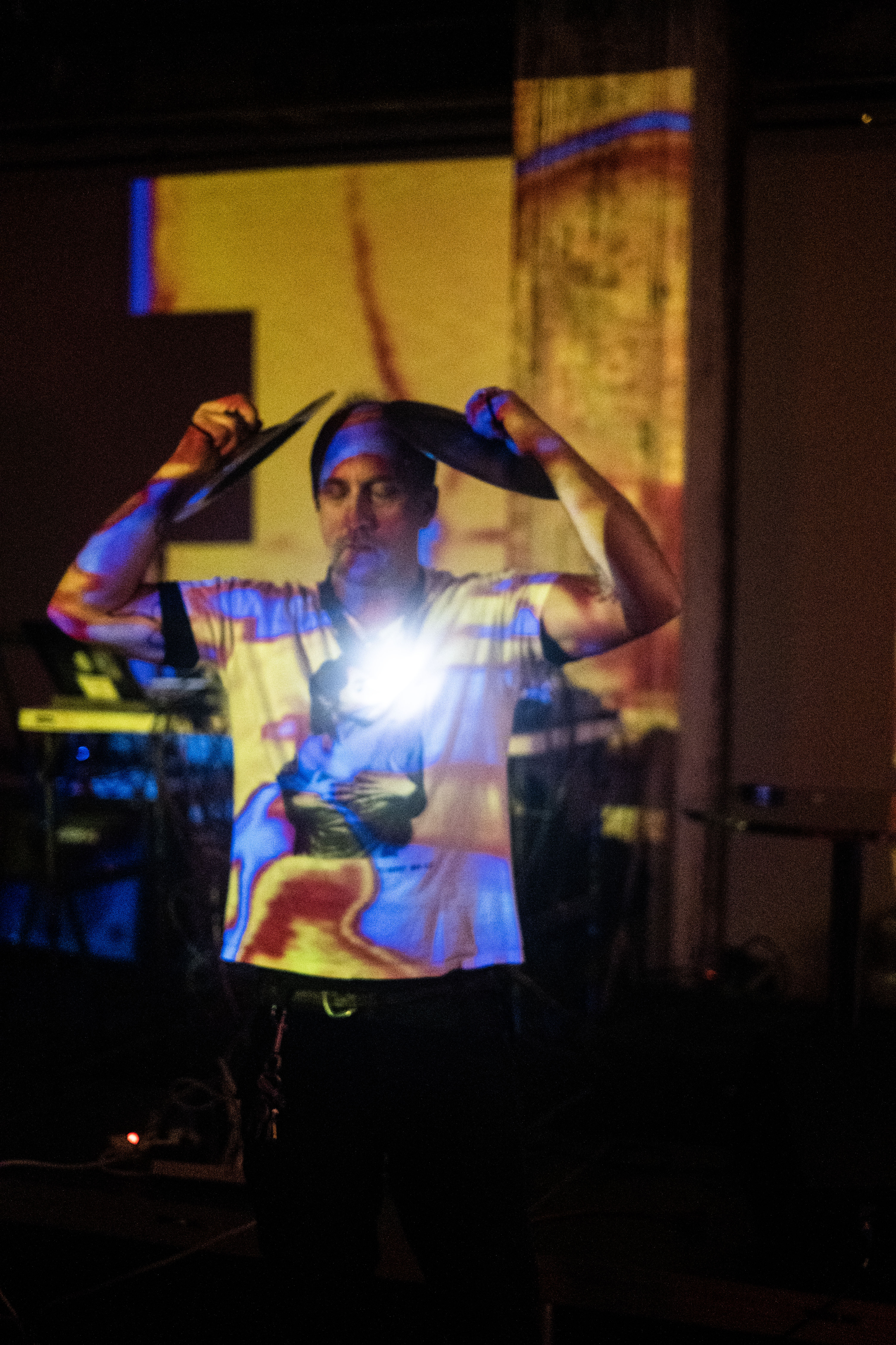 A figure in a white graphic T-shirt has their eyes closed, while they stand and hold two cymbals over their head. Yellow and blue projections are cast onto them on stage.