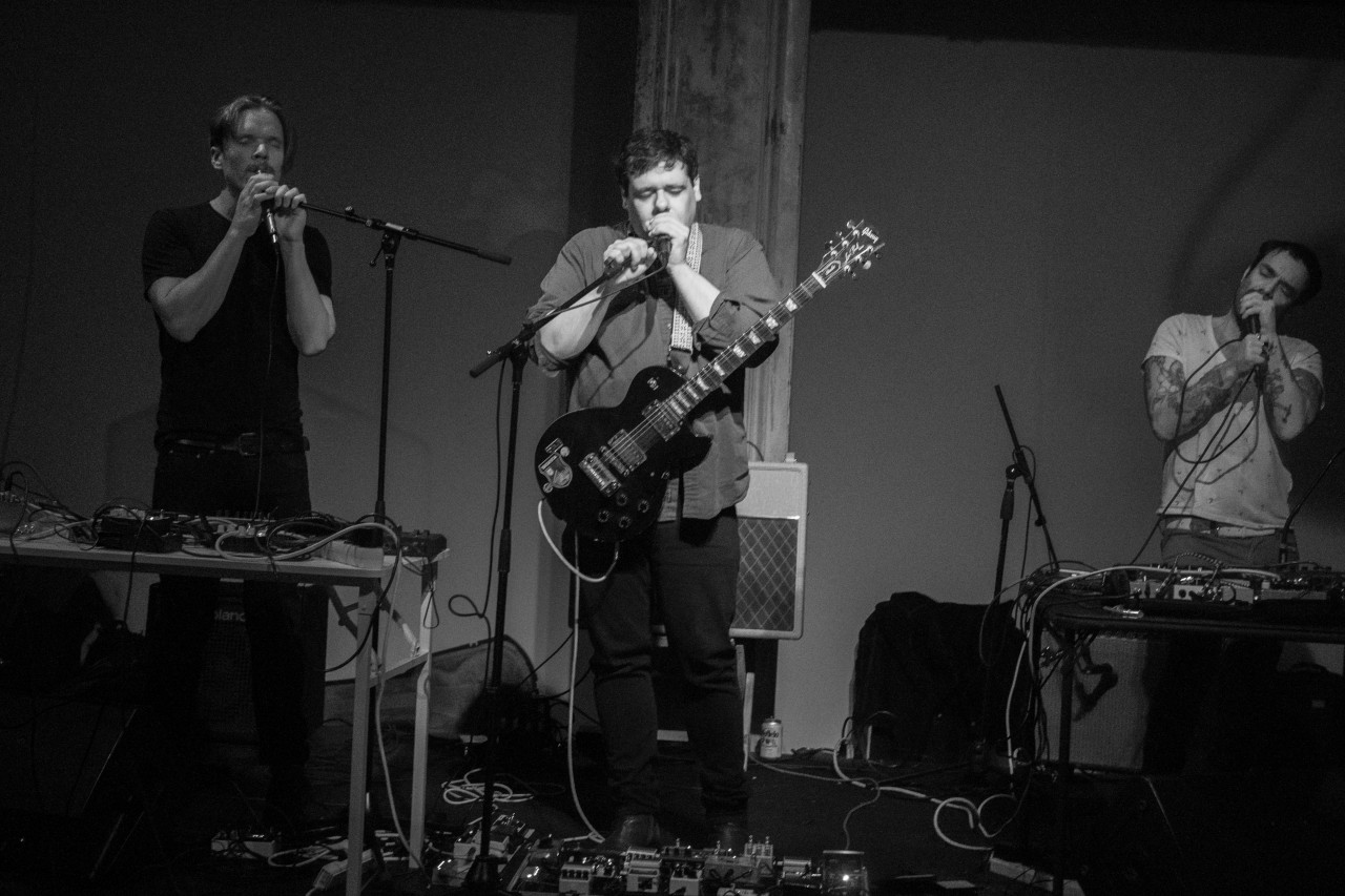 Black-and-white image of three figures standing and performing on stage. Each figure holds a microphone to their face, their eyes closed in concentration. The figure in the middle is wearing a guitar while the other two on the left and right stand behind tables full of audio equipment.