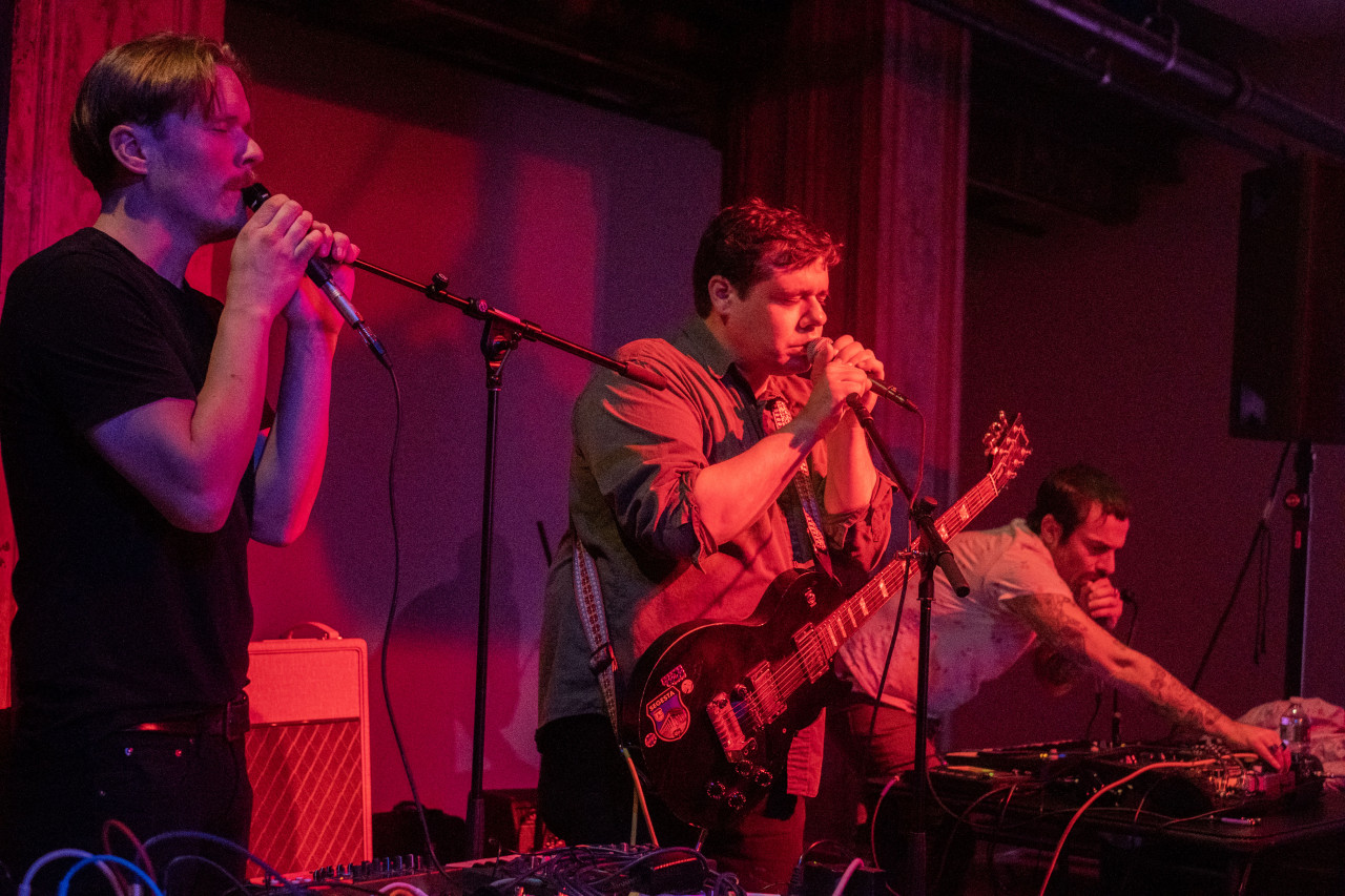 Three figures stand on a stage cast in red and purple light from a projector. They each hold a microphone to their face, their eyes closed in concentration. The figure in the middle wears a guitar, and the figure all the way on the right leans forward on a table with audio equipment.