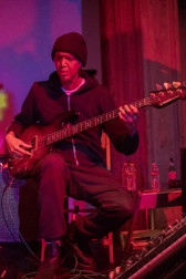 A seated figure in a black hoodie and black beanie plays the guitar with red and purple light cast onto them.