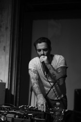 Black-and-white image of a figure performing on stage. They hold a microphone to their mouth and the other hand plays a synthesizer.