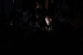 Color image of an individual seated and intently looking into a computer screen. The bright glow from the computer illuminates the performer's face and the electronic gear on the worktable before him.
