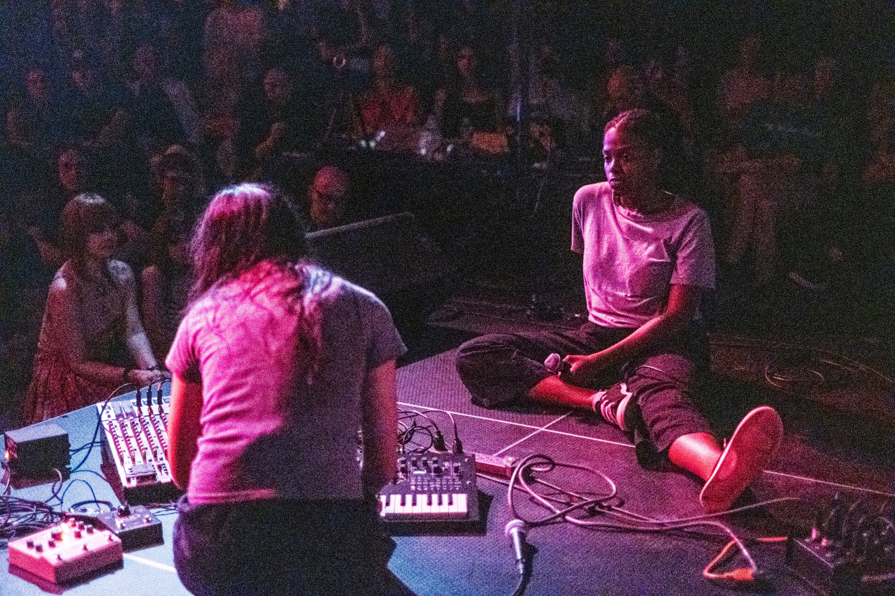 A grainy color image of two performers facing each other on a stage, with a large audience observing in a dark room. One individual on stage is sitting and faces the camera, watching her performance partner. The other individual has their back turned to the camera, as they crouch over musical equipment on the stage floor. A pinkish-red hue from some spotlights illuminates the on-stage performers.