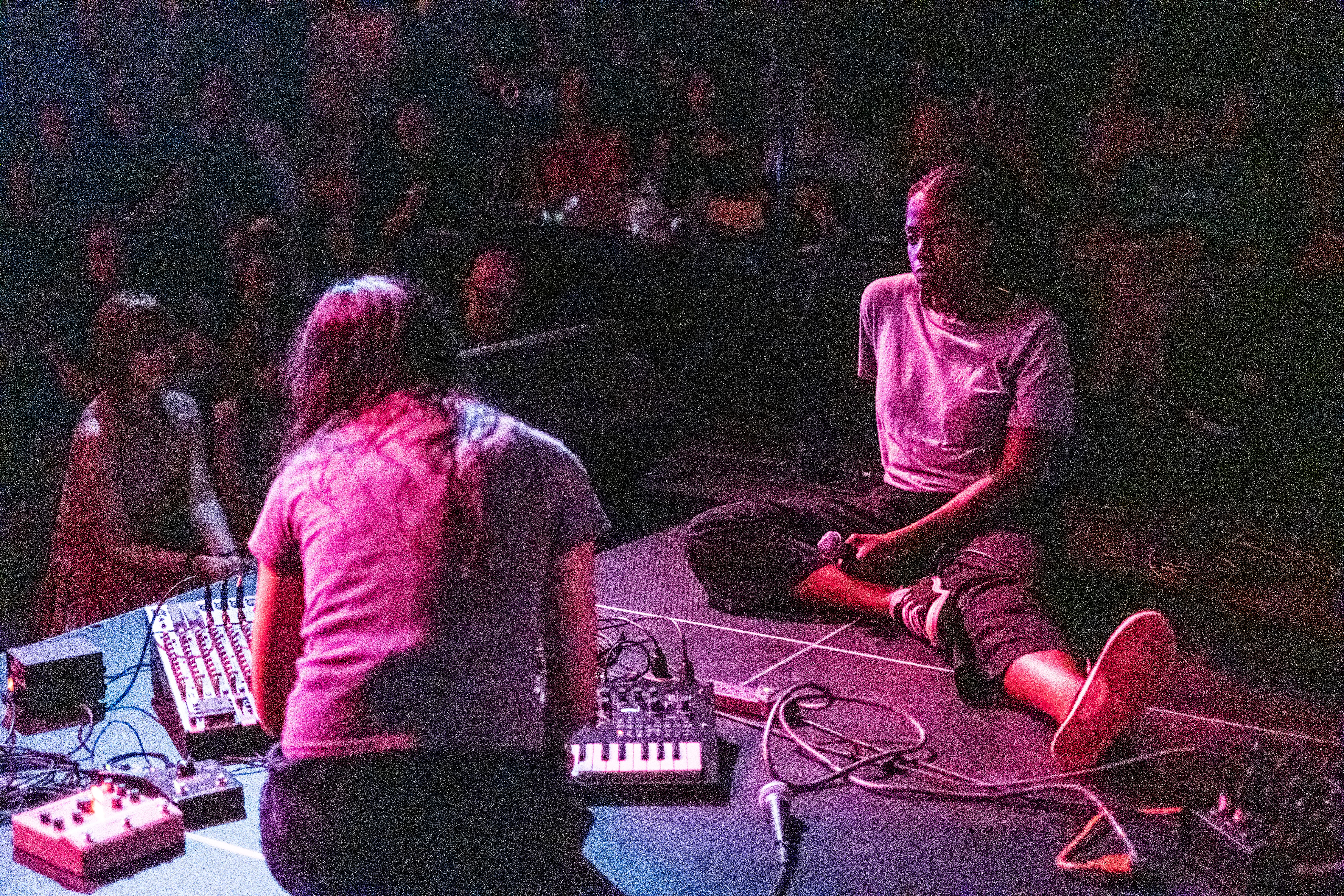 A grainy color image of two performers facing each other on a stage, with a large audience observing in a dark room. One individual on stage is sitting and faces the camera, watching her performance partner. The other individual has their back turned to the camera, as they crouch over musical equipment on the stage floor. A pinkish-red hue from some spotlights illuminates the on-stage performers.
