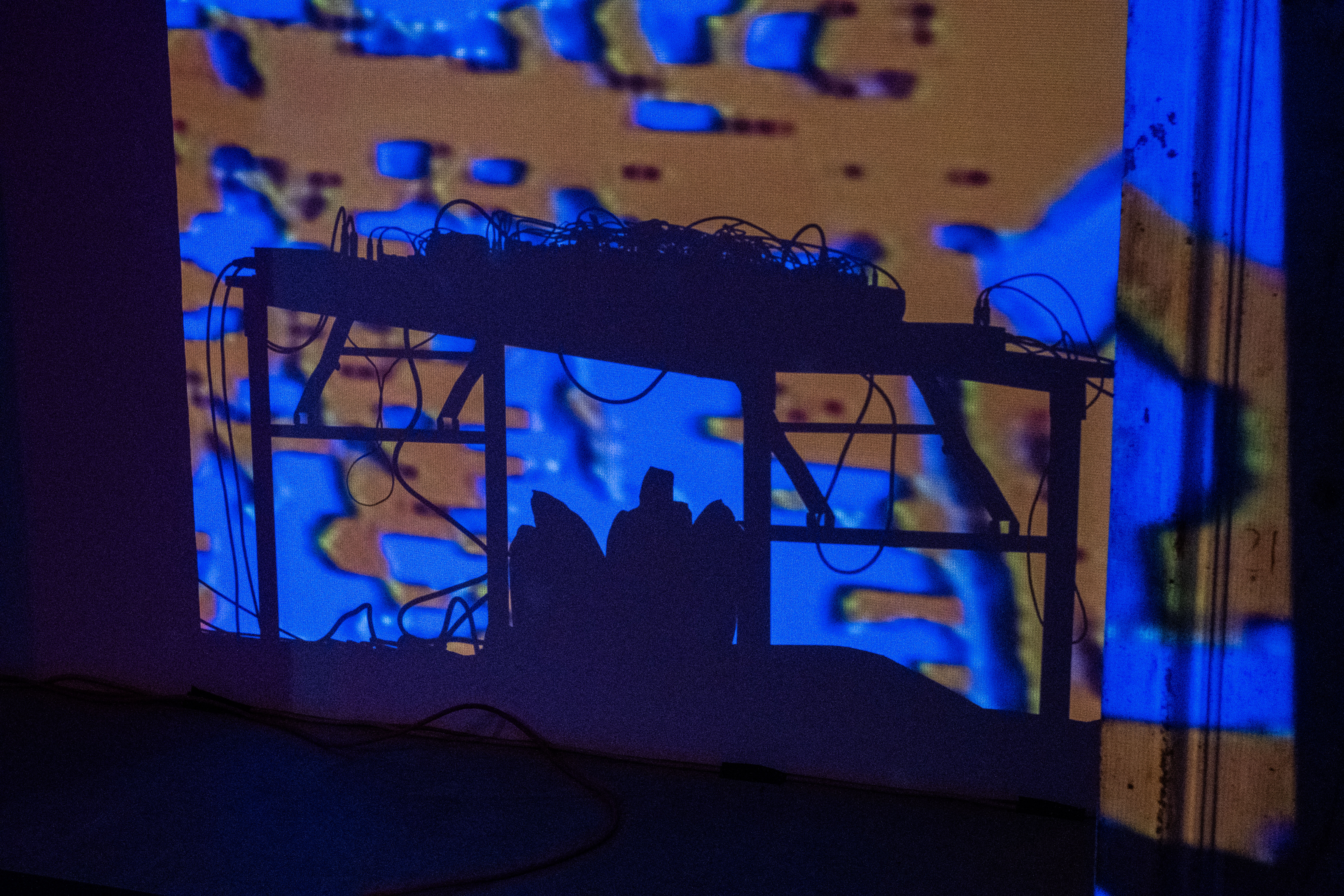 Color image of a dark room, illuminated only by a projection of blue and orange amorphous shapes on a wall. The shadow of a table with dj equipment and gear is cast against the colorful projected image.