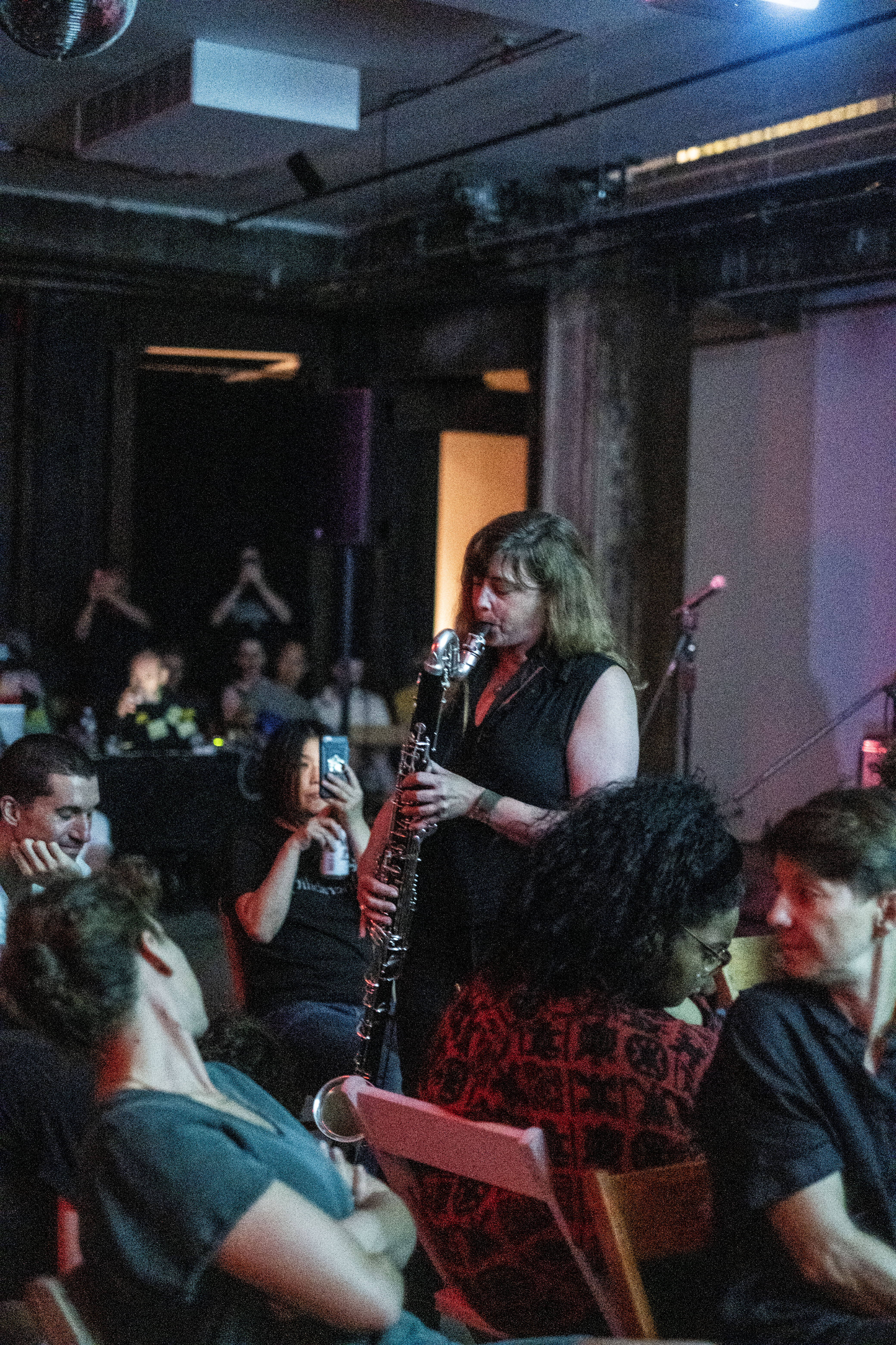 Color image of a performer captured in the middle of a bassoon performance. The musician stands in the middle of a crowd of seated guests who are observing her in a dimly lit performance venue.