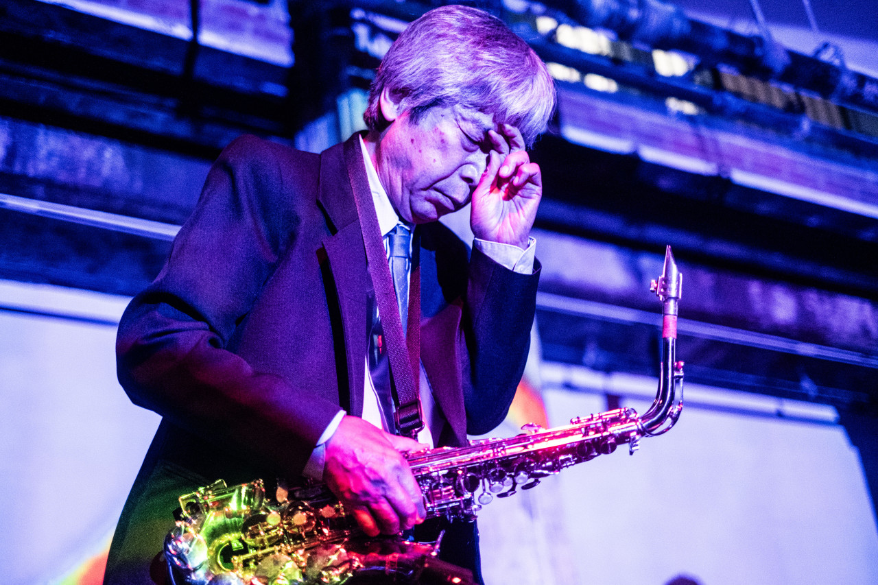 Close-up color image of a musician holding a saxaphone to his body. One hand holds the instrument while the other hand is held up to his face in a contemplative gesture. Hues from diffused rainbow and bright purple lights are cast against the performer and in the interior space.