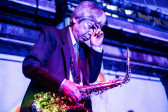 Close-up color image of a musician holding a saxaphone to his body. One hand holds the instrument while the other hand is held up to his face in a contemplative gesture. Hues from diffused rainbow and bright purple lights are cast against the performer and in the interior space.