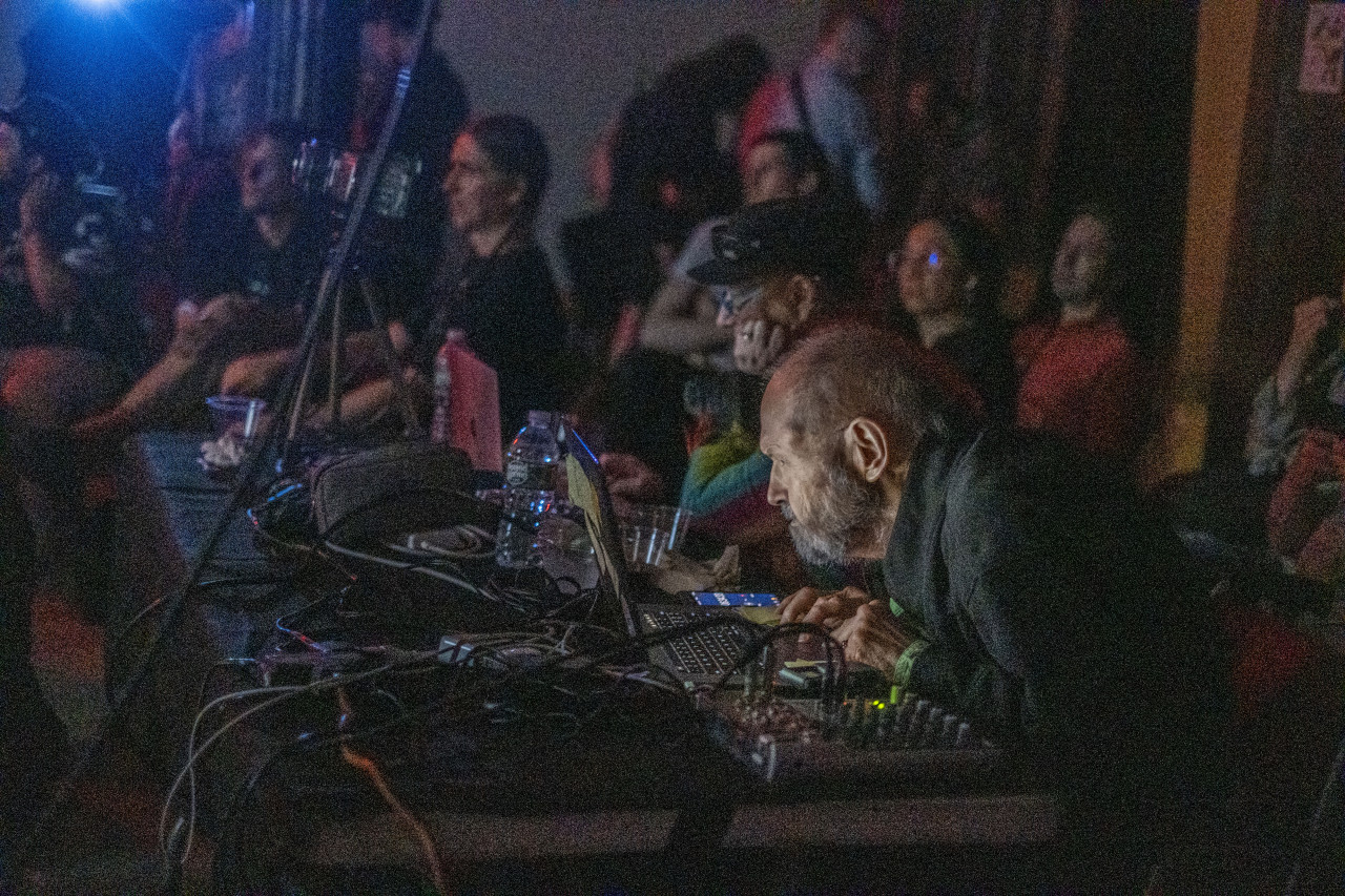 Color image of an individual seated at a table with a variety of musical and electronic equipment and gear in front of him. The individual stares with focus at a computer screen, which illuminates his face in the darkened performance space. A variety of guests can been seen seated all around the musician