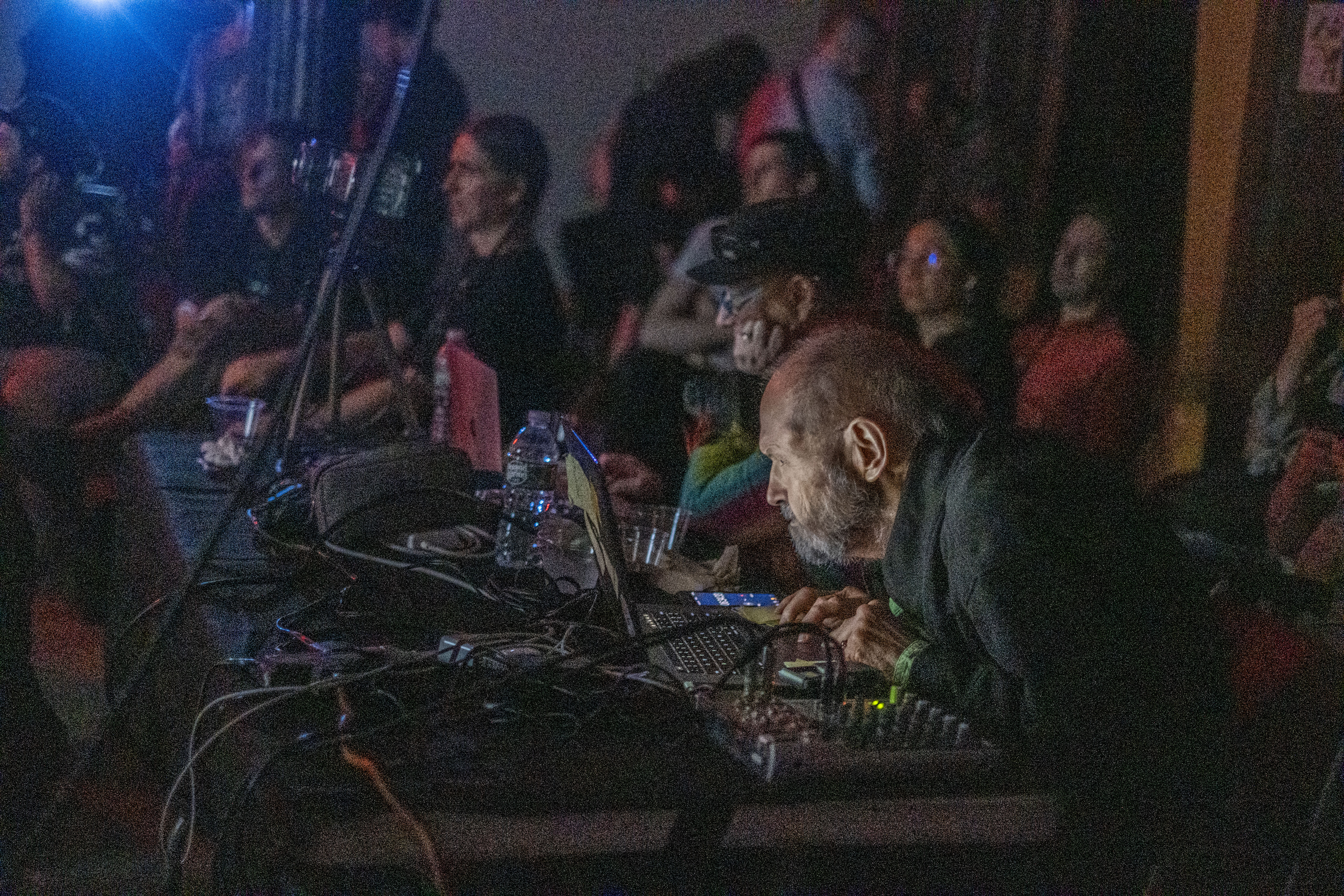 Color image of an individual seated at a table with a variety of musical and electronic equipment and gear in front of him. The individual stares with focus at a computer screen, which illuminates his face in the darkened performance space. A variety of guests can been seen seated all around the musician's work table.