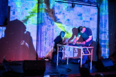 Color image of an individual captured midst performing at a table set up with DJ equipment. Another performer is seen behind him, playing the saxaphone in a dark performance space illuminated only by bright blue, pink and neon yellow projections. A large shadow of a person taking a photo with a camera can be seen cast on the projection wall to the left of the image.