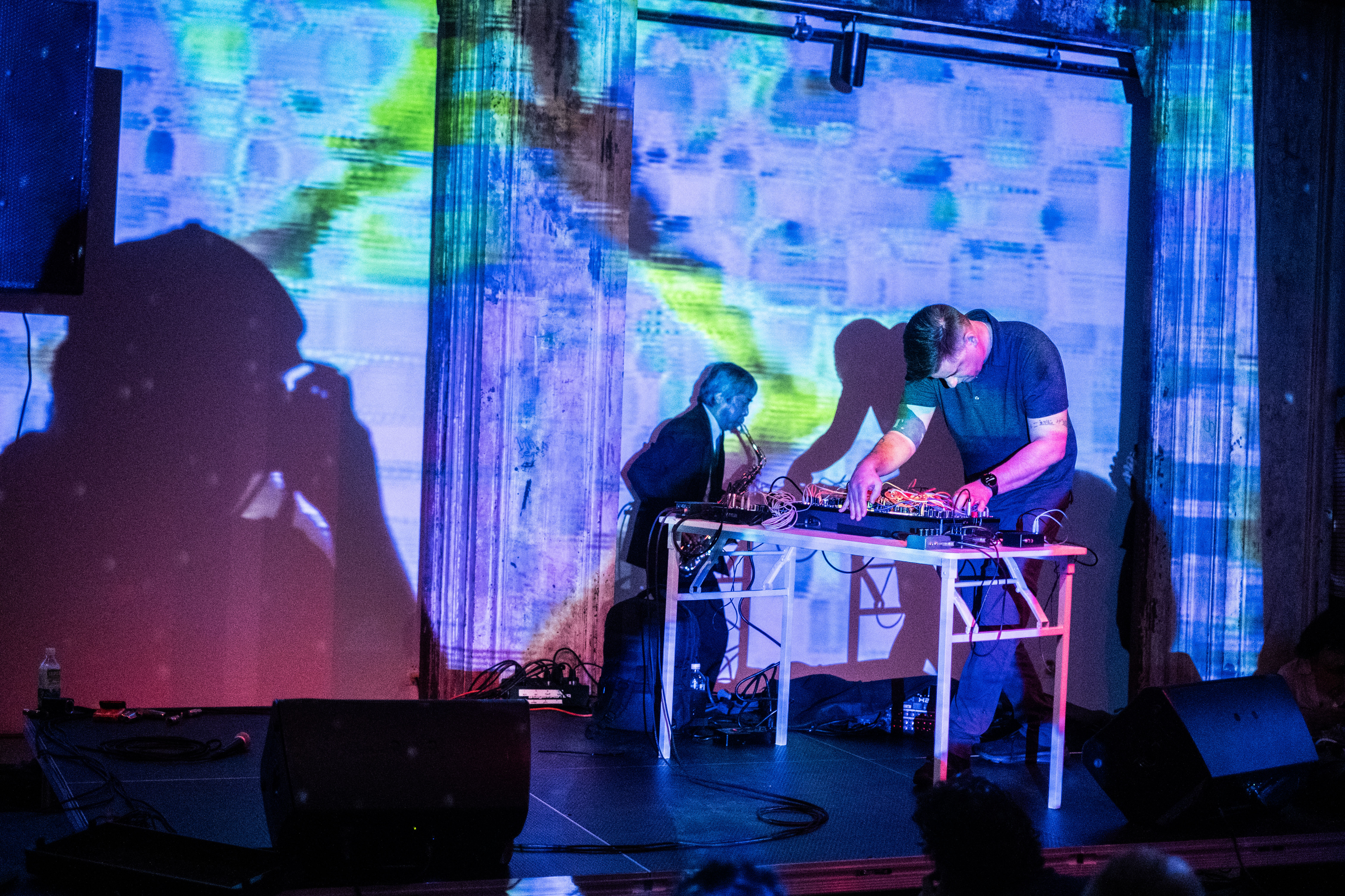 Color image of an individual captured midst performing at a table set up with DJ equipment. Another performer is seen behind him, playing the saxaphone in a dark performance space illuminated only by bright blue, pink and neon yellow projections. A large shadow of a person taking a photo with a camera can be seen cast on the projection wall to the left of the image.