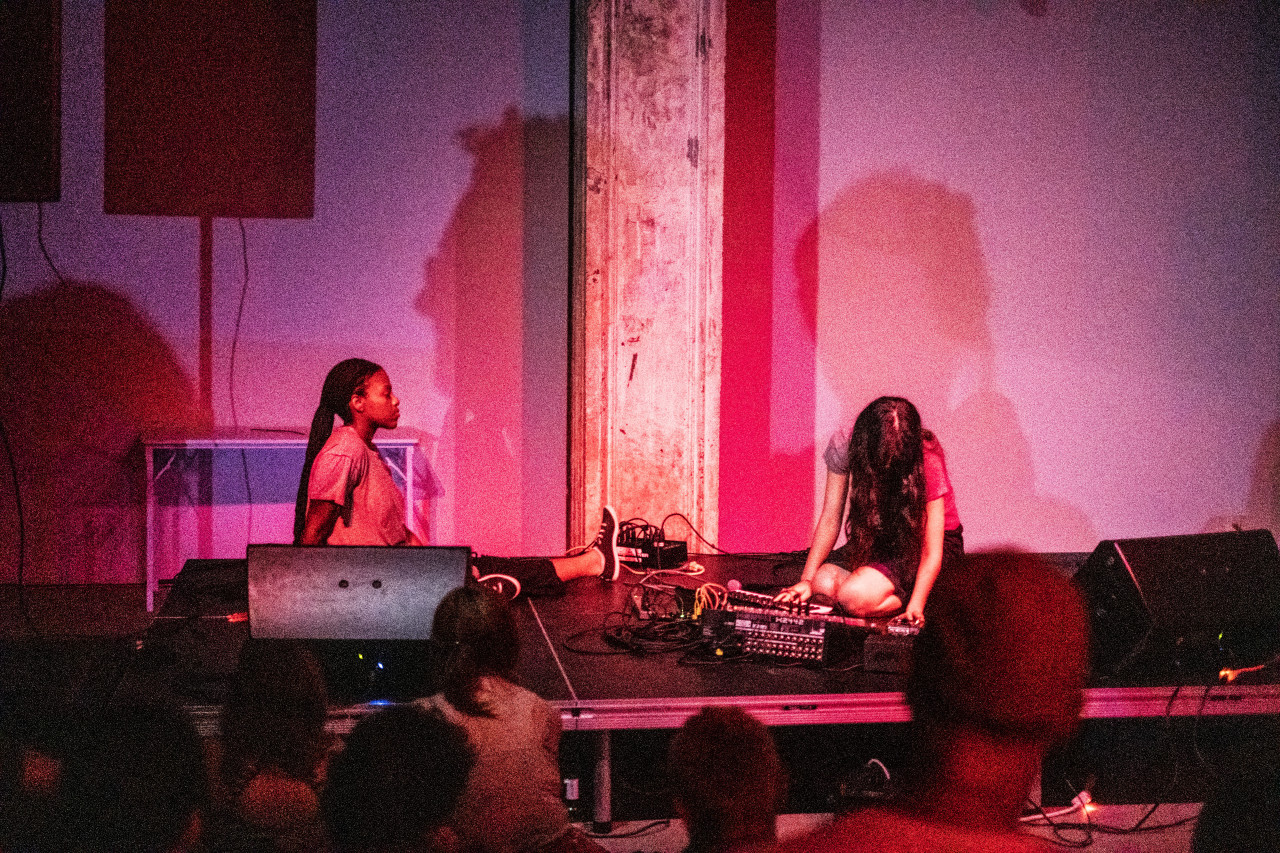 Color image of two performers sitting on a stage, with a variety of musical gear between them. The backs of heads from audience members observing the performers can be seen at the bottom of the image. A pinkish-red spotlight illuminates the dark performance space and the individuals on the stage.
