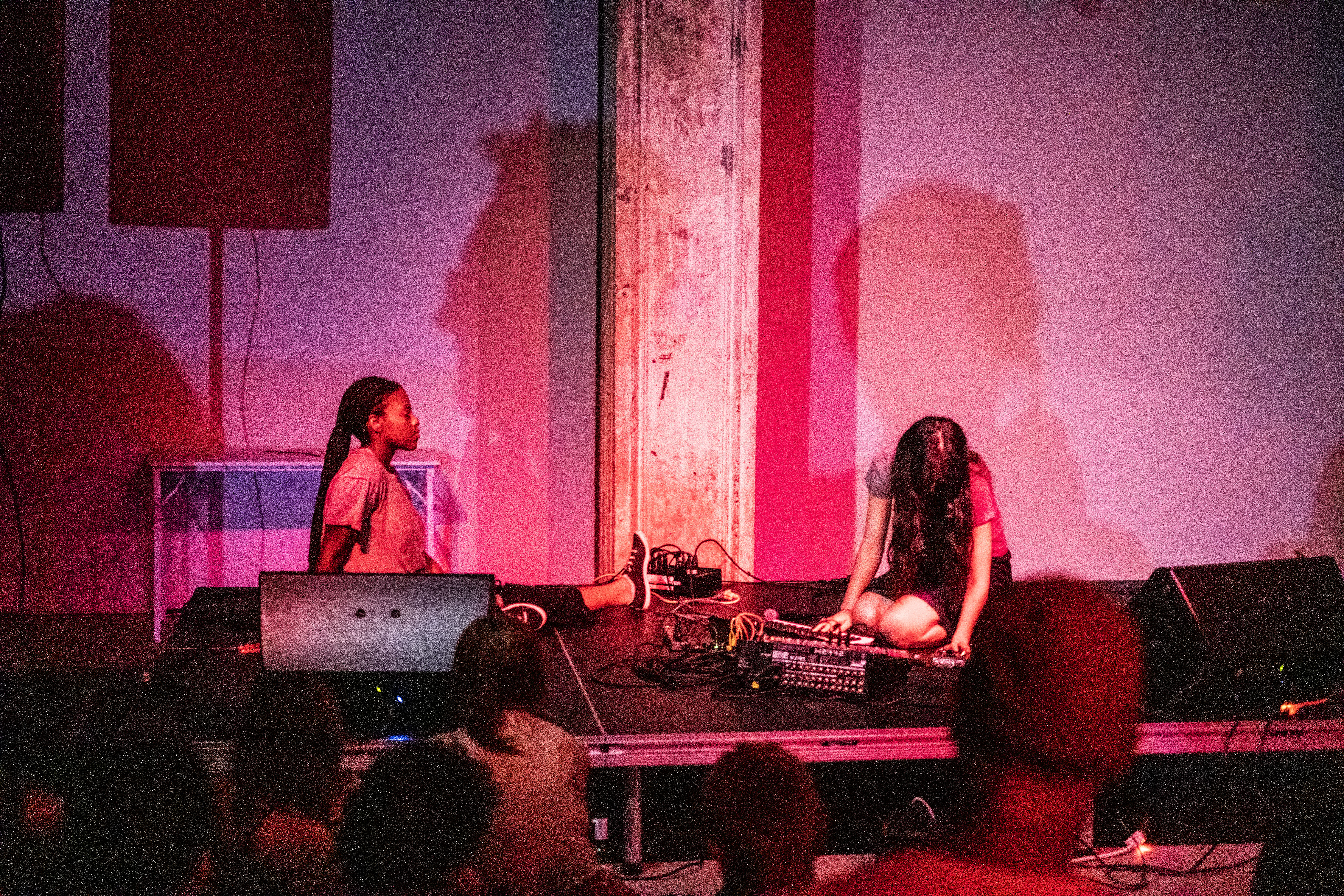 Color image of two performers sitting on a stage, with a variety of musical gear between them. The backs of heads from audience members observing the performers can be seen at the bottom of the image. A pinkish-red spotlight illuminates the dark performance space and the individuals on the stage.