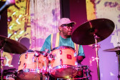 A close-up color image of a drummer captured in the middle of a performance. Abstracted projections of bright yellow and purple hues are cast against the musician, who looks out at something out of view.