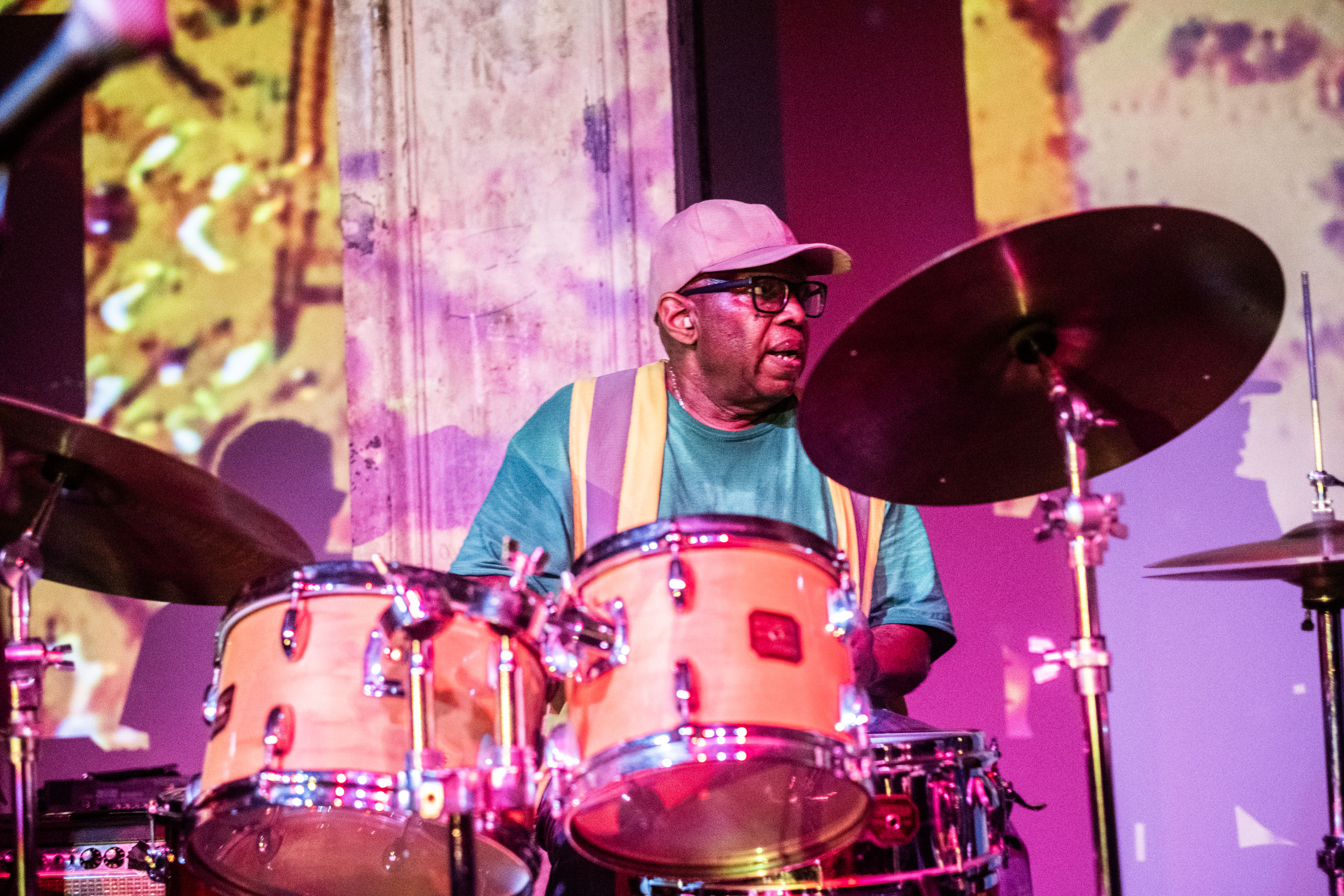 A close-up color image of a drummer captured in the middle of a performance. Abstracted projections of bright yellow and purple hues are cast against the musician, who looks out at something out of view.