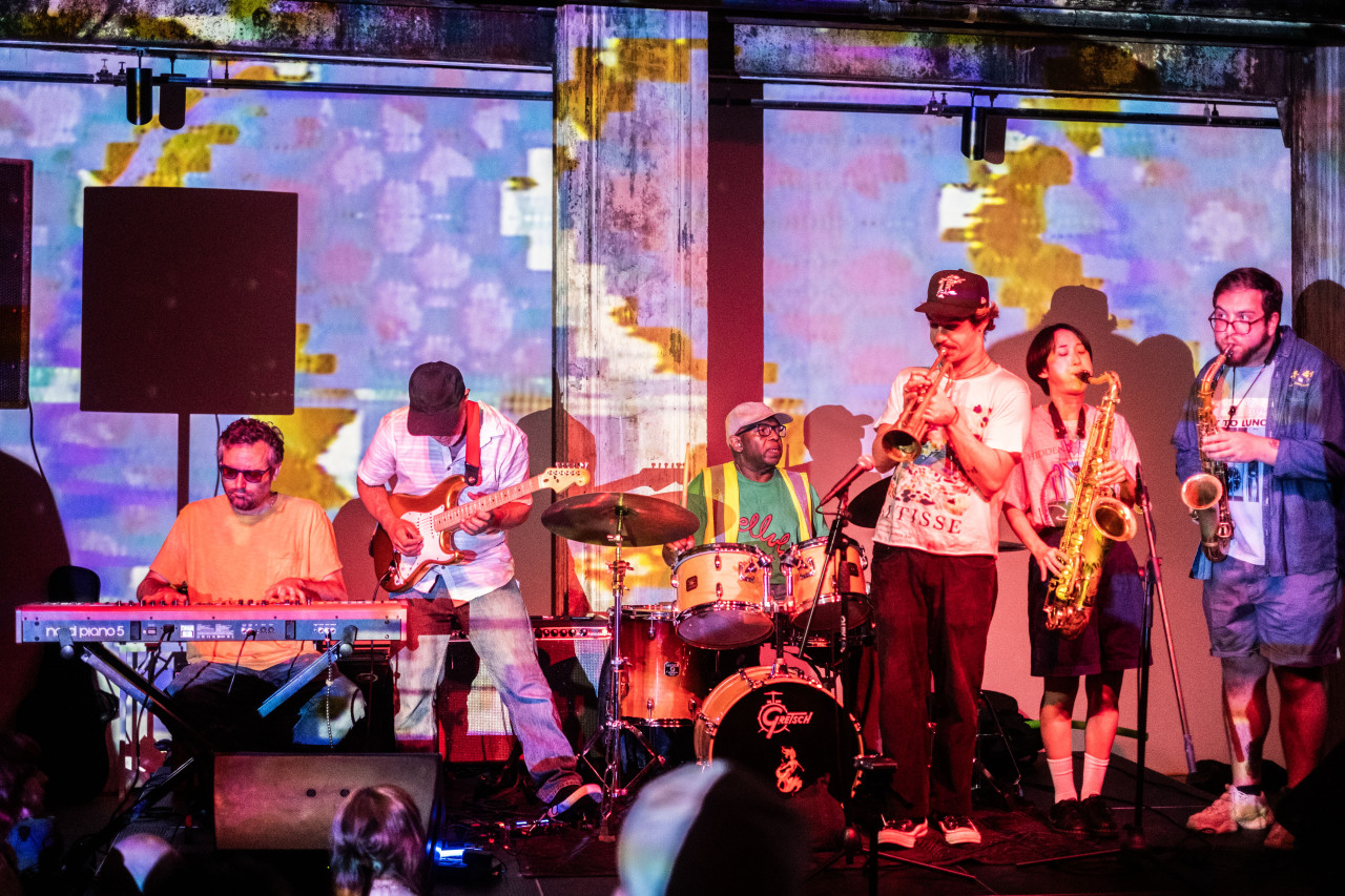Color image of a band of performers playing music on a stage together, colorful and patterned projections illuminate the group and the wall behind them. In order: group includes a keyboardist, a bass player, a drummer, a trumpet player and two saxaphone players.