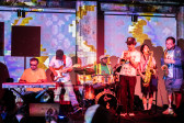 Color image of a band of performers playing music on a stage together, colorful and patterned projections illuminate the group and the wall behind them. In order: group includes a keyboardist, a bass player, a drummer, a trumpet player and two saxaphone players.