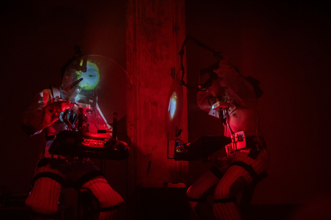 Color photo of two performers kneeling on the floor of a dark performance venue. A deep red glowing light emanating from between the performers serves as the only source of illumination. The crouching individuals are wearing quilted pants and have a variety of nightvision gear attached to their legs and torsos.