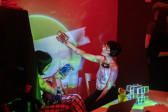 Color photo of two performers sitting on the ground in a dark venue space that is illuminated by deep red overhead lights. The performers are holding up clear, geometric objects against a neon yellow and blue projection on a wall.
