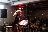 Color photo of a woman captured mid-performance in a dimly lit venue. The performer is situated before a microphone and a music stand, while a crowd of seated and standing audience members attentively watch. Bright spotted lights from a disco ball out of view sparsely illuminate the environment.