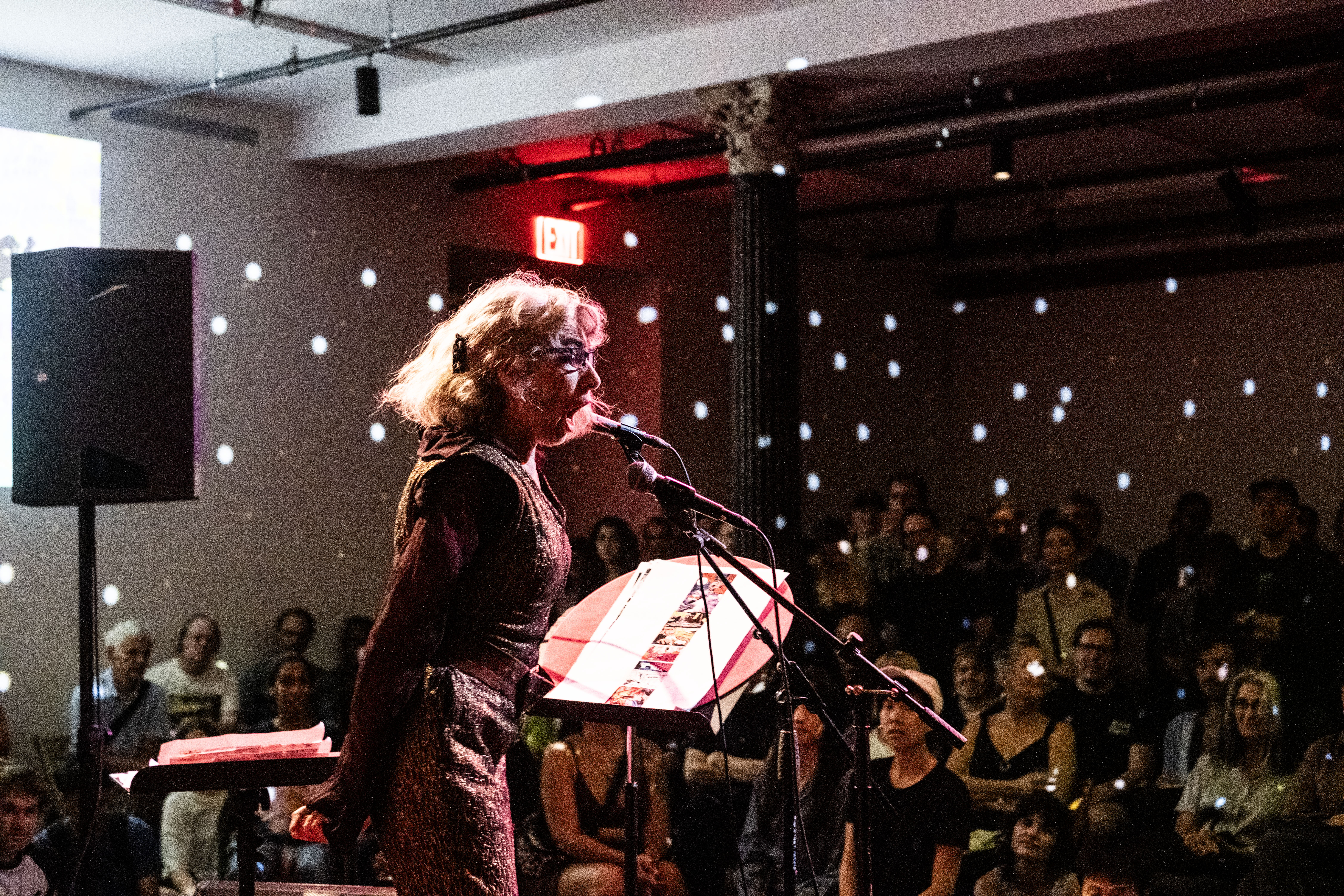 Color photo of a woman captured mid-performance in a dimly lit venue. The performer is situated before a microphone and a music stand, while a crowd of seated and standing audience members attentively watch. Bright spotted lights from a disco ball out of view sparsely illuminate the environment.