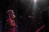 Color photo of a woman captured with her eyes closed, standing before a pair of microphones in a darkened performance space. Blue spotted lights and the reddish glow from a different light source shine onto the performer.
