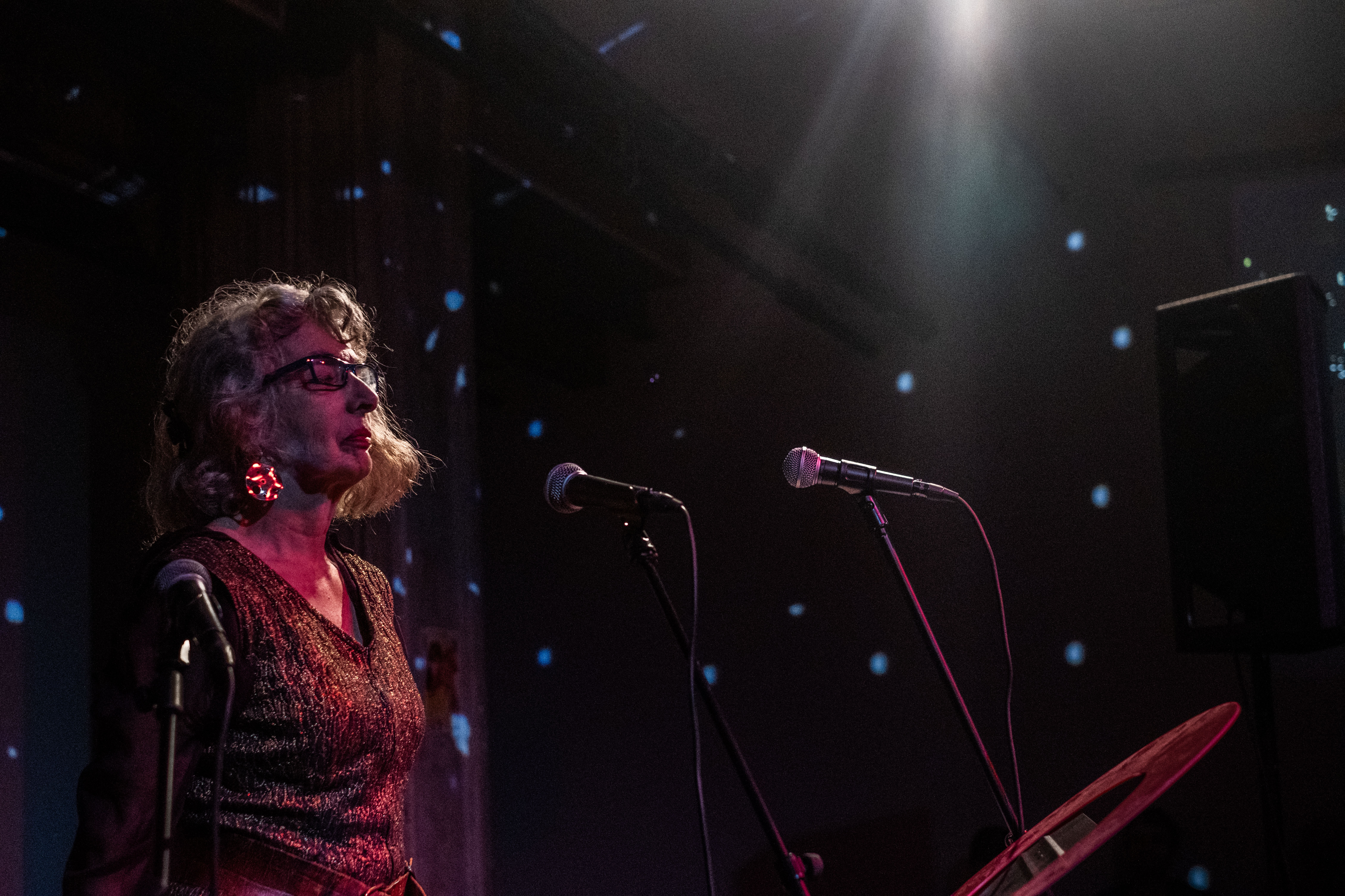 Color photo of a woman captured with her eyes closed, standing before a pair of microphones in a darkened performance space. Blue spotted lights and the reddish glow from a different light source shine onto the performer.