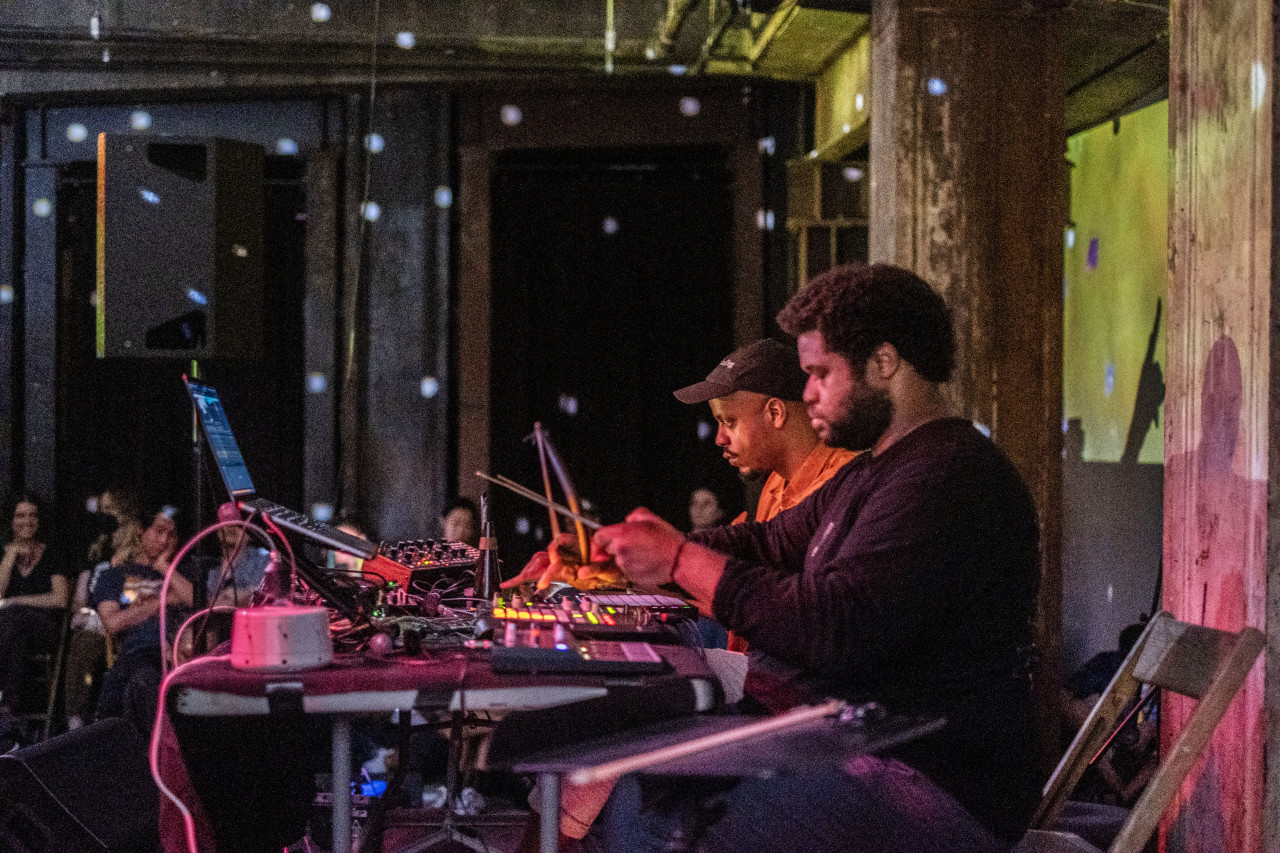 Color photo of two men sitting at a worktable that holds various musical and electronic equipment. In the background, various audience members sit and observe the performers within a dimly lit space. Yellow projections shine on the wall behind the performers along with blue and pink spotted lights.