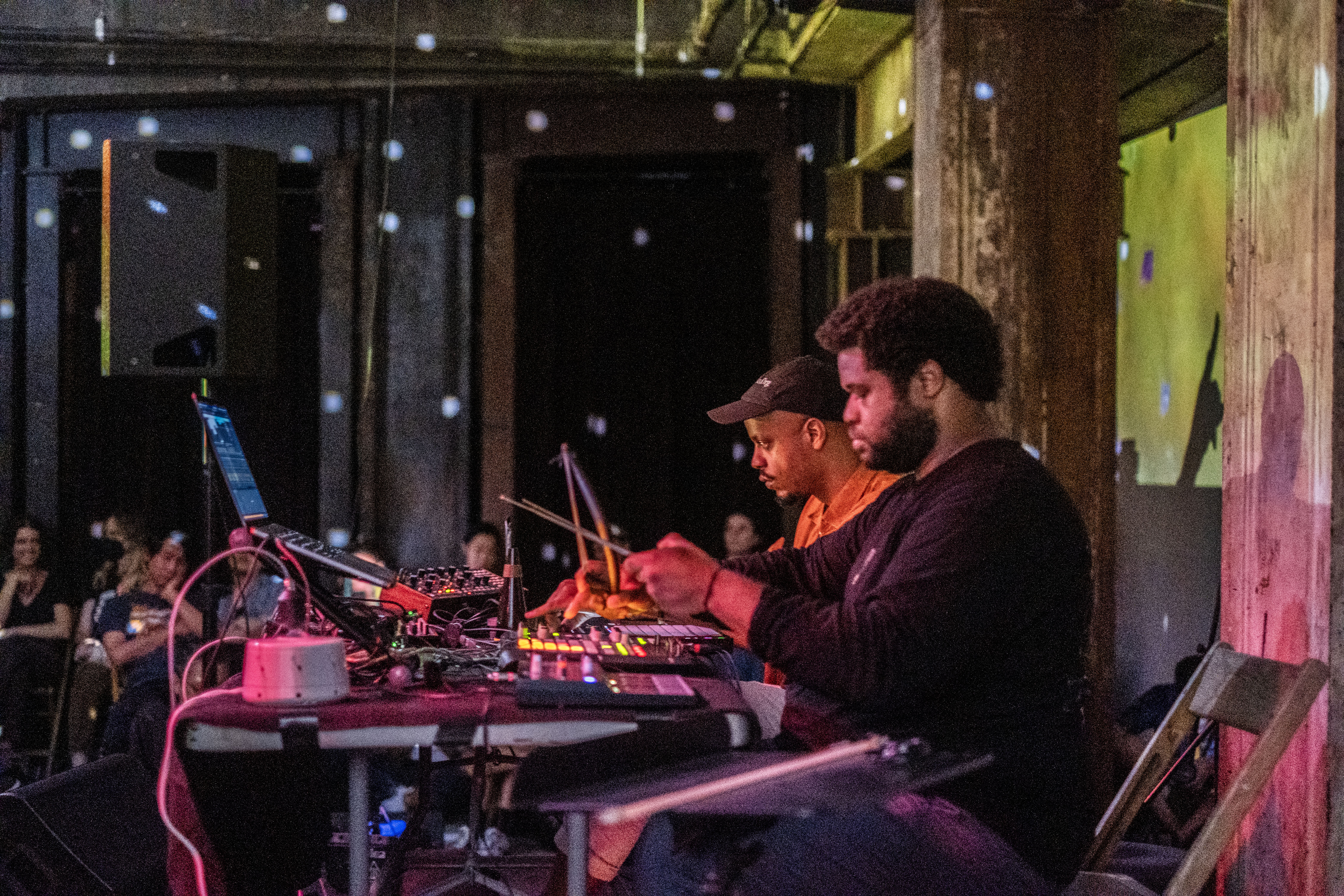 Color photo of two men sitting at a worktable that holds various musical and electronic equipment. In the background, various audience members sit and observe the performers within a dimly lit space. Yellow projections shine on the wall behind the performers along with blue and pink spotted lights.