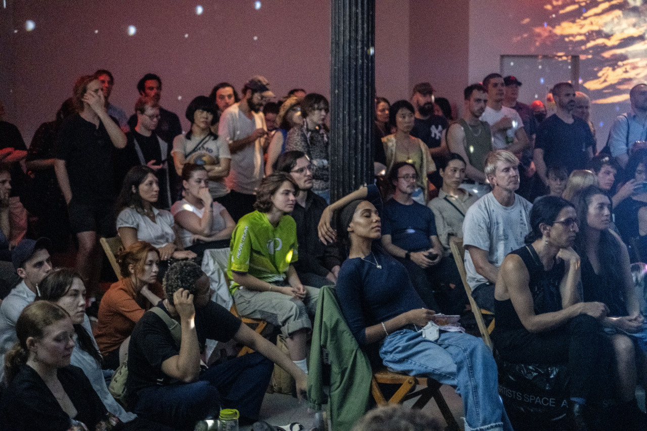 Color photo of a large gathering of audience members seated and standing in a dimly lit performance space. The venue is illuminated by white spotted lights from a discoball out of view and soft, pink projections.