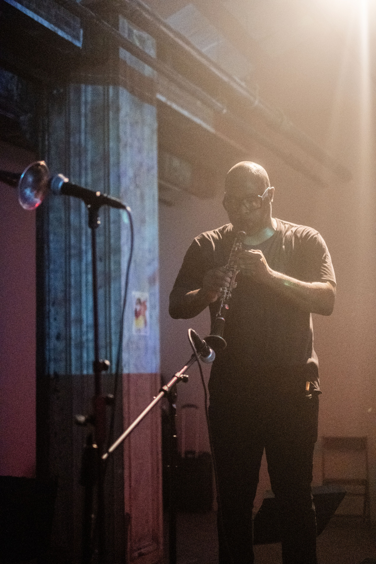 Color photo of a musician captured mid-performance as he plays his clarinet into a microphone positioned near his instrument. The musician stands in a dim venue space with a bright, warm spotlight that shines directly onto him.