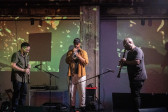 Color photo of three musicians captured mid-performance on stage in a low-lit venue space. The central musician plays the trumpet, and is flanked at either side by men playing clarinets. Abstract and fragmented projections in green and yellow-orange shine onto the musicians and cast onto the wall behind them.