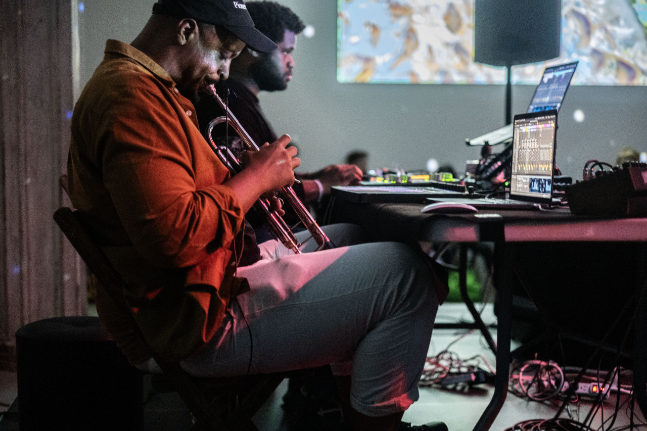 Color photo of two men sitting at a work table that holds various electronic and musical equipment. The man seated closest to the camera is playing a trumpet, while the other performer at his side actively works at a computer. The performance venue is dimly lit while a projection and some pink overhead lights provide some illumination.