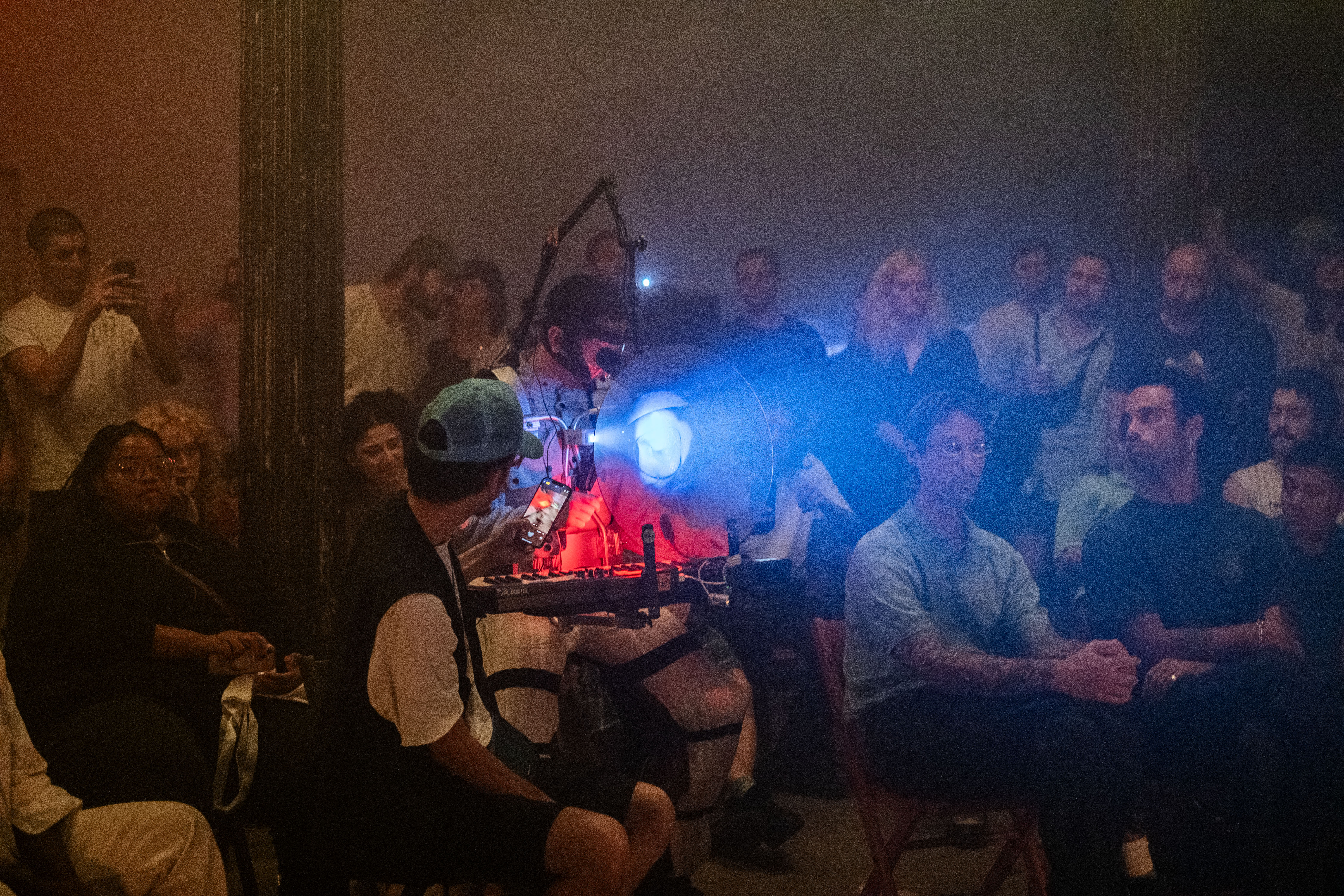 Color photo of a performer wearing a quilted suit with a variety of equipment strapped to his body. A bright blue glowing projection emantes from the performer's gear. He is surrounded by seated audience members who observe the performance in a dimly lit venue.