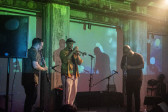 Color photo of three musicians captured mid-performance on stage in a dark venue space. The performer in the middle plays the trumpet and is flanked at either side by two men playing clarinets. The musicians are illuminated by green and blue abstract projections that are cast on the wall behind them.