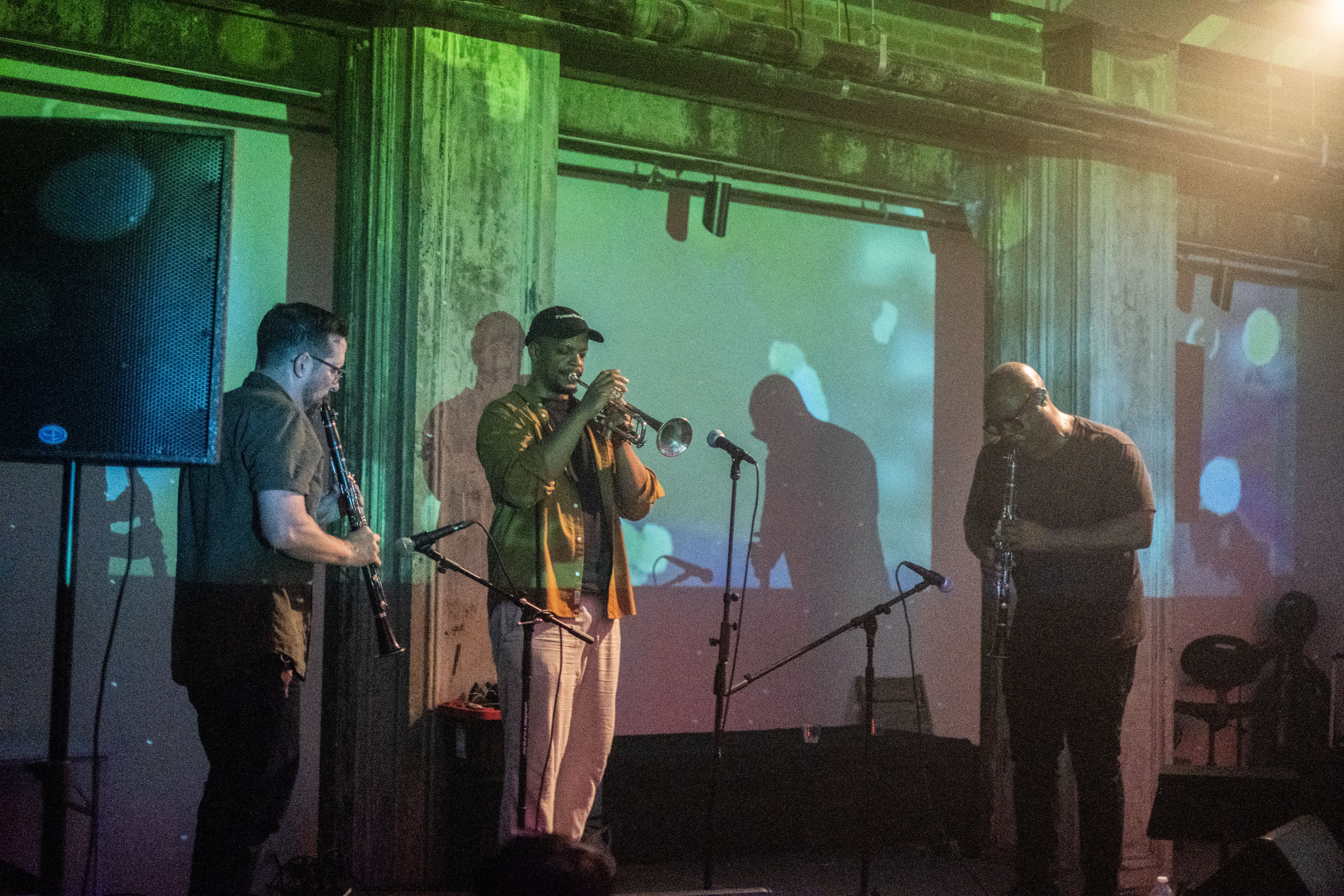 Color photo of three musicians captured mid-performance on stage in a dark venue space. The performer in the middle plays the trumpet and is flanked at either side by two men playing clarinets. The musicians are illuminated by green and blue abstract projections that are cast on the wall behind them.