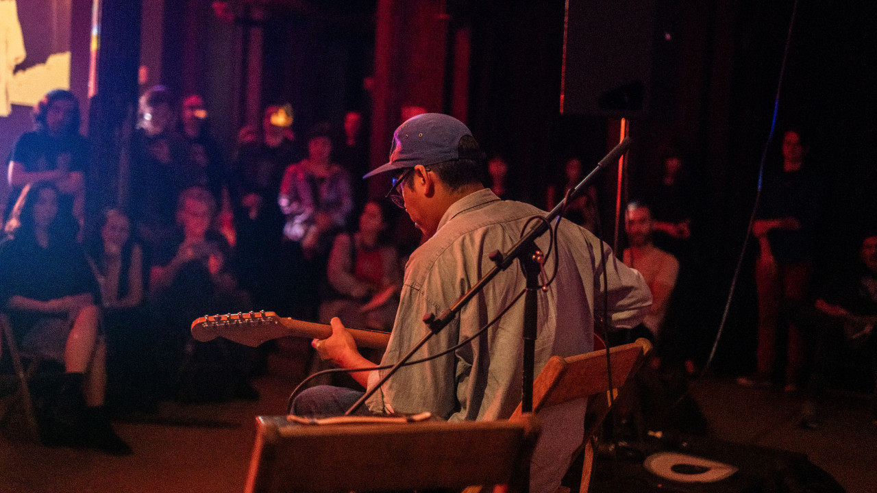 A color image of a figure, seen from behind, performing on stage. The figure sits on a chair and plays the guitar, and the audience is on the background. Red, and purple light fills the room.