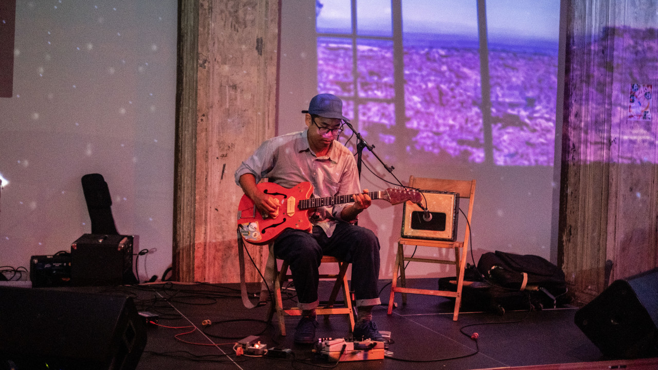 A figure performs on the stage. He is sitting on a chair playing a guitar. To the right, there is another chair with an amp on the seat. Nature images are projected onto the stage, and back wall.
