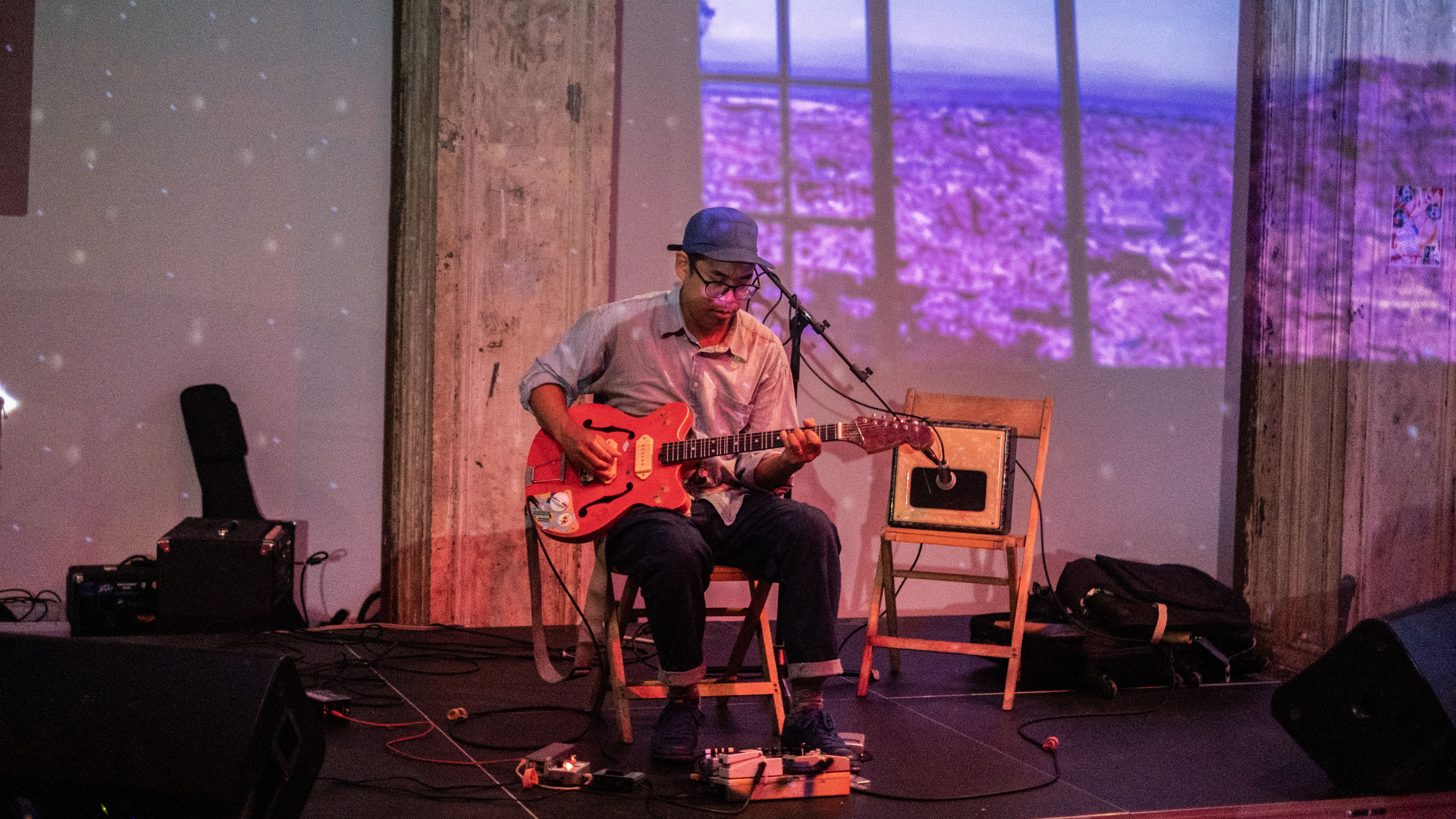 A figure performs on the stage. He is sitting on a chair playing a guitar. To the right, there is another chair with an amp on the seat. Nature images are projected onto the stage, and back wall.