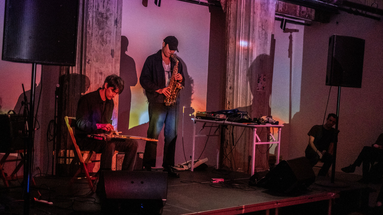 Two musicians perform on stage. To the left, one of them sits on a chair with an electric guitar laying on his legs. To the right, a musician stands playing the saxophone. Rainbow lights are projected onto the stage.