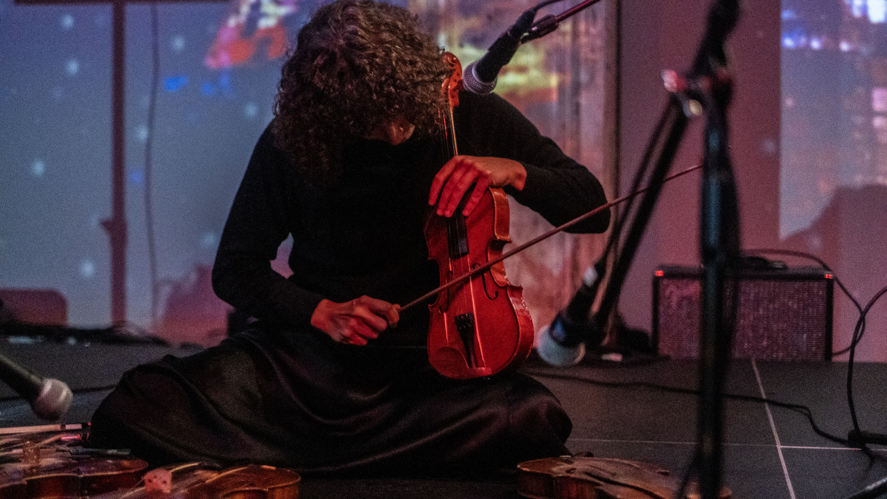 A close-up image of a musician surrounded by three microphones is sitting on the floor playing the violin. Three other violins lay on the stage before her. Abstract images are projected onto the back.