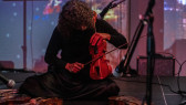 A close-up image of a musician surrounded by three microphones is sitting on the floor playing the violin. Three other violins lay on the stage before her. Abstract images are projected onto the back.