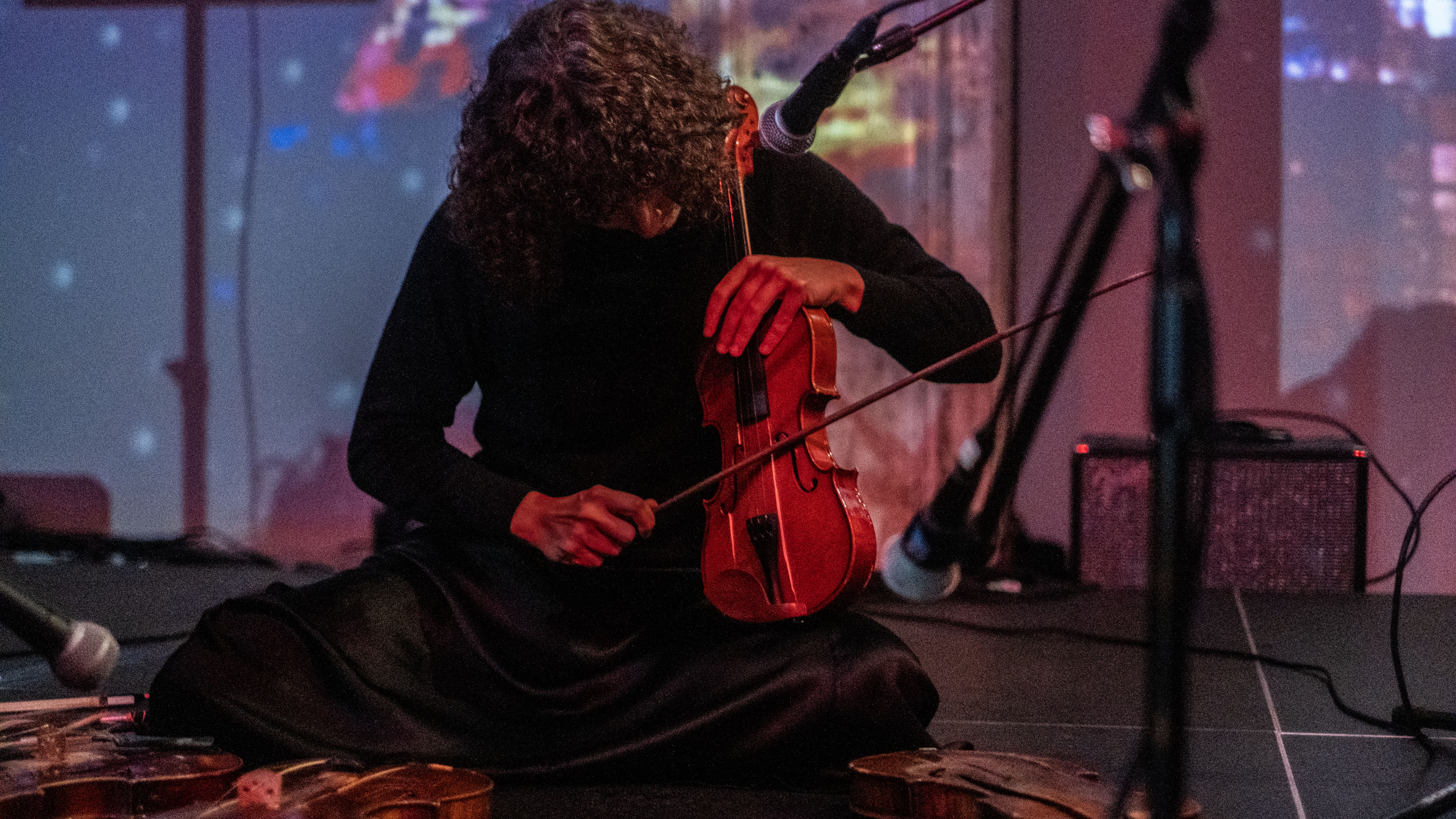 A close-up image of a musician surrounded by three microphones is sitting on the floor playing the violin. Three other violins lay on the stage before her. Abstract images are projected onto the back.