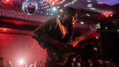 A close-up icolor mage of a musician playing the guitar. Red light cast onto them. A disco ball hangs from the ceiling, projecting white dots onto the room.