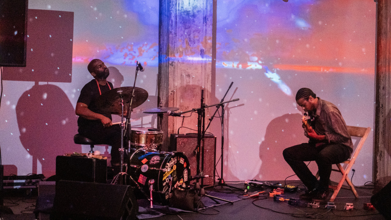Two figures musicians perform on stage. On the left, a figure is playing the drums. On the right, a guitar player, sits on a chair.  Blue and red lights are projected onto the stage.