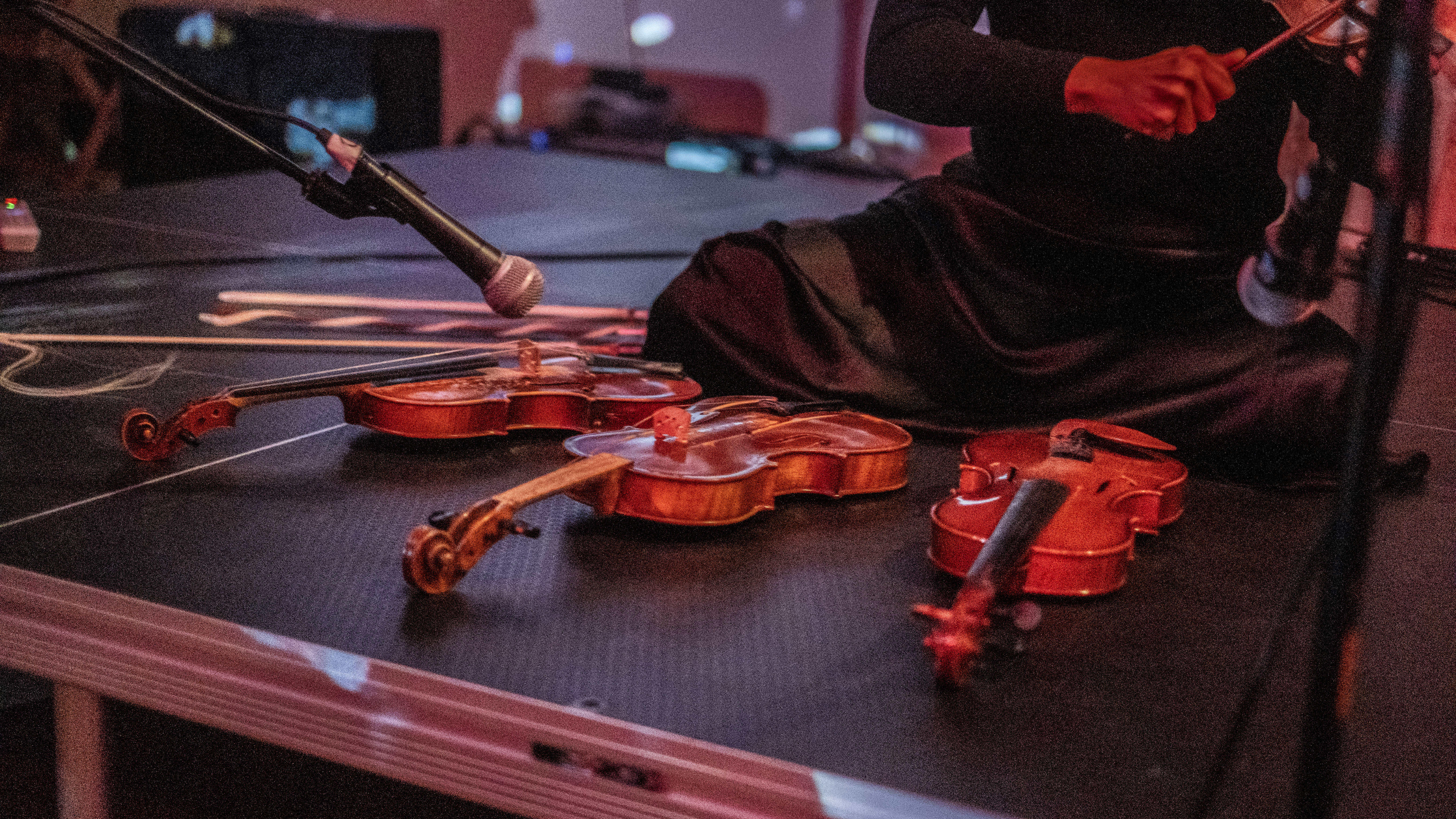 A close-up image of a musician sitting on the floor playing the violin. Three other violins lay on the stage before her.
